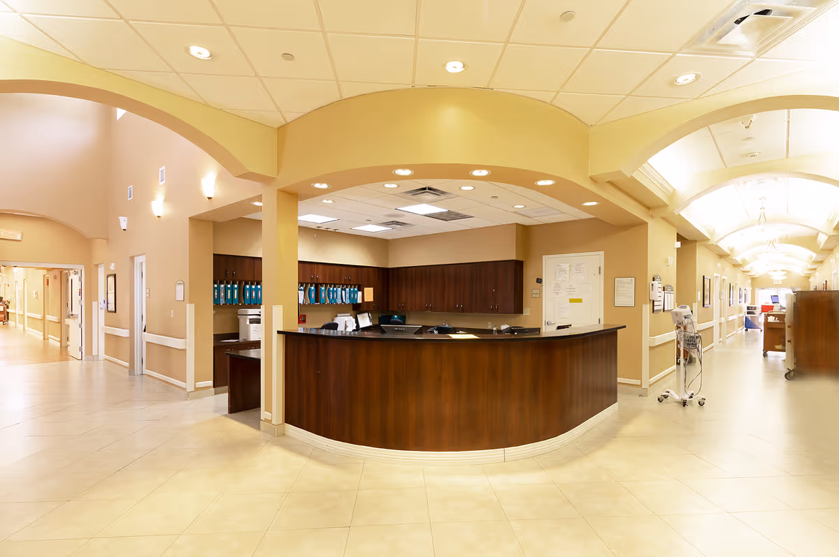 A spacious reception or nurse station area inside a senior living facility with a curved wooden desk, overhead recessed lighting, beige walls, and tiled floors. The area is flanked by two long hallways with handrails and additional lighting fixtures on the ceiling.