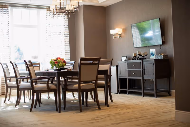 A bright dining room with a rectangular wooden table surrounded by eight cushioned chairs. A floral centerpiece is placed on the table. Large windows with sheer curtains allow natural light to fill the room. On the right wall, there is a mounted flat-screen TV above a dark wooden sideboard with decorative items and candles. The room has beige walls and light-colored flooring.