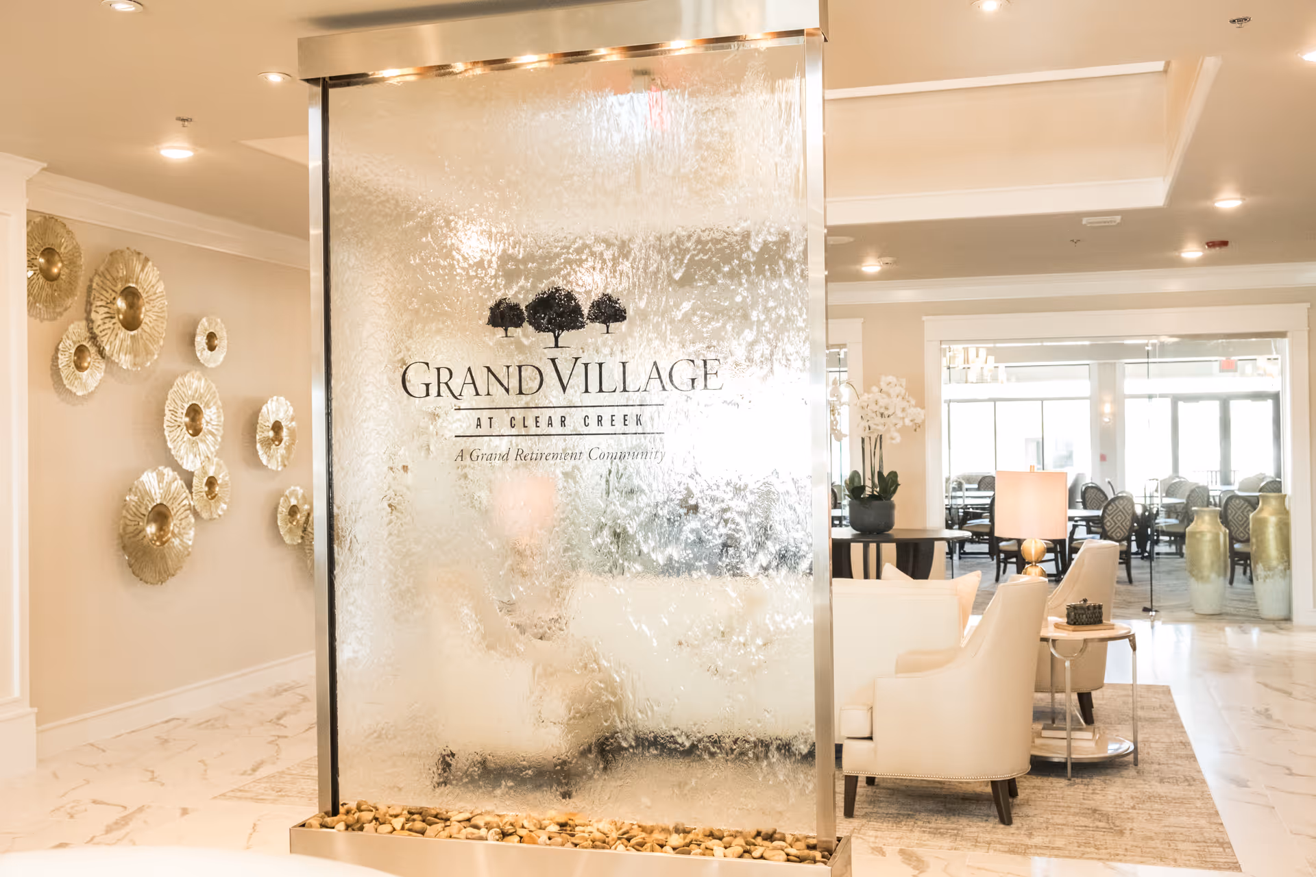 Bright lobby seating area with a large indoor glass water wall displaying the 'Grand Village at Clear Creek' logo and decorative furniture.