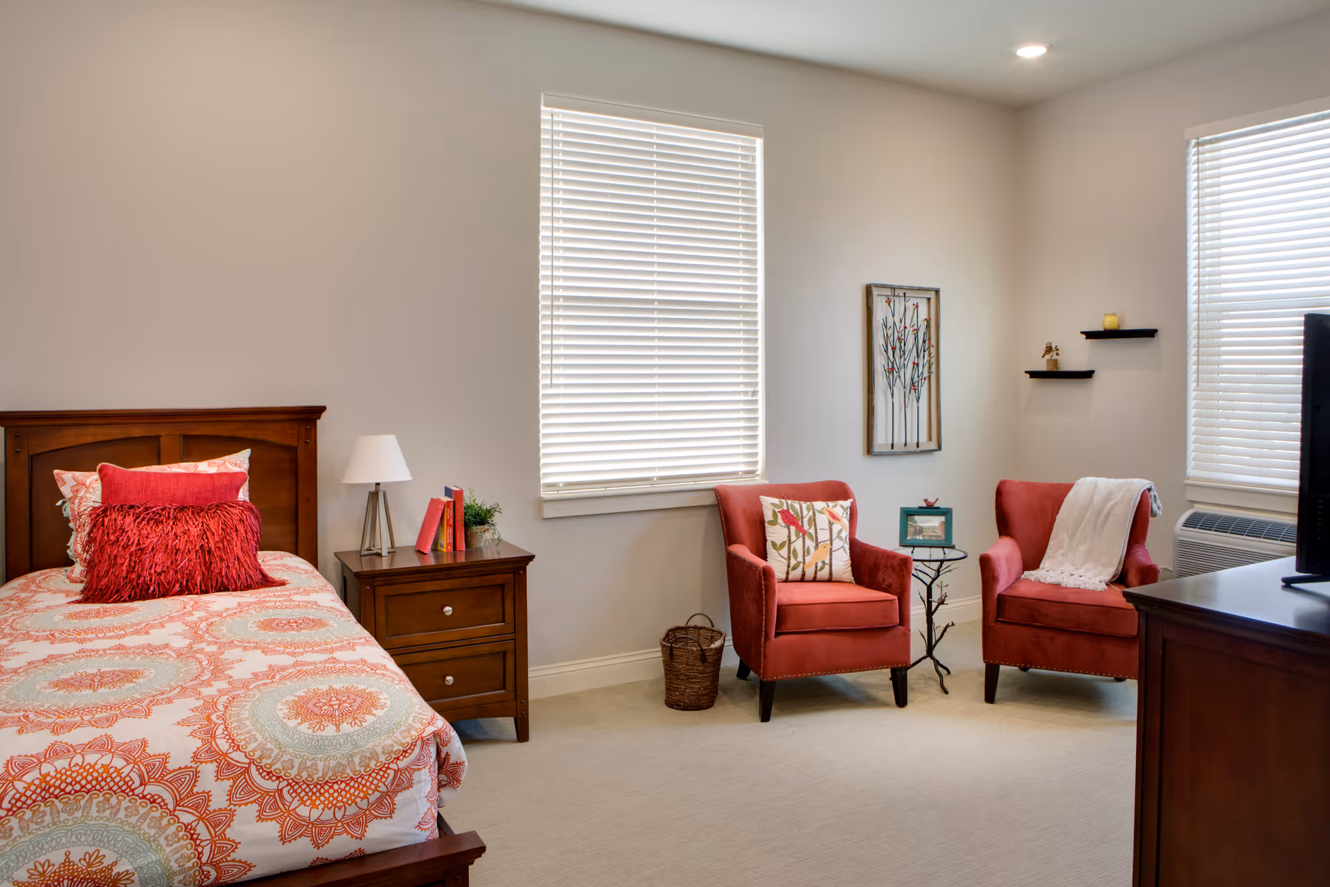 A cozy bedroom with a single bed featuring a colorful patterned bedspread and red pillows. Next to the bed is a wooden nightstand with a lamp, books, and a small plant. Two red armchairs with decorative pillows and a white throw blanket are positioned near two windows with blinds. A small round table sits between the chairs, and there is wall art and small shelves on the wall. A wooden dresser with a TV on top is partially visible on the right side.