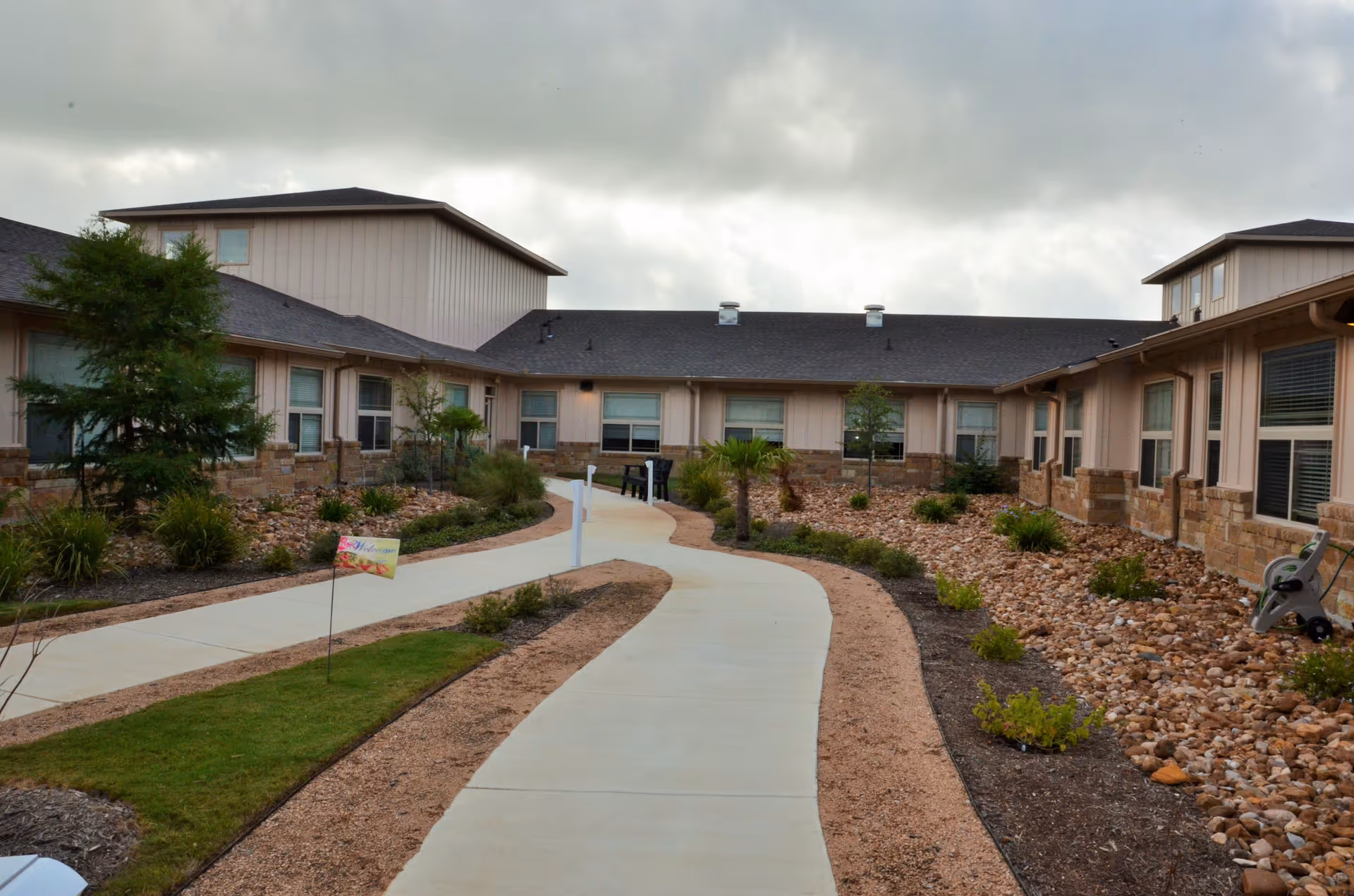 Outdoor courtyard area of The Grandview of Westover Hills with a winding concrete pathway, landscaped with small bushes, rocks, and trees, surrounded by a single-story building under a cloudy sky.