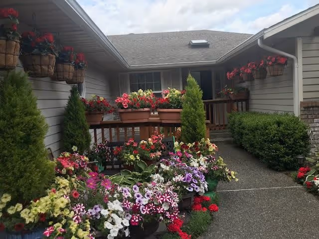 Outdoor garden area at a residential care facility with a variety of colorful flowers in pots and planters along a pathway leading to the entrance of a building with gray siding and a shingled roof.