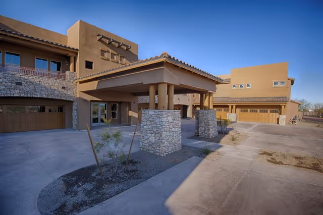Front entrance of a southwestern-style building with a covered porte-cochère supported by stone pillars and attached garage bays under a clear blue sky.