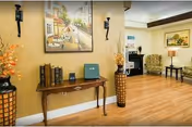 Interior view of a senior living facility common area with wooden flooring, a wooden console table holding books and a small safe, two tall decorative vases with flowers on either side, wall sconces, and framed artwork on the walls. In the background, there are upholstered chairs and a small table with a lamp.