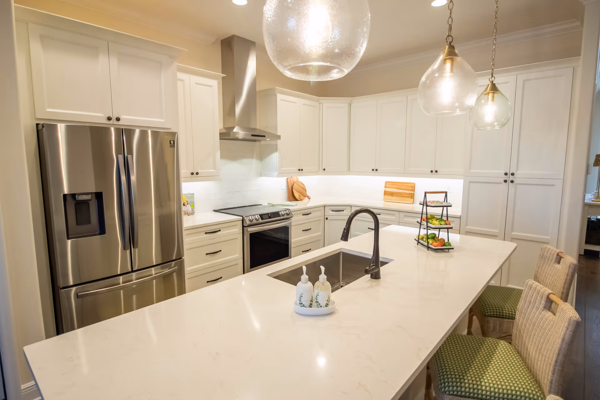 Modern kitchen with white cabinetry, stainless steel refrigerator and oven, a large white marble island with a sink and faucet, three glass pendant lights hanging above the island, and two wicker chairs with green cushions.