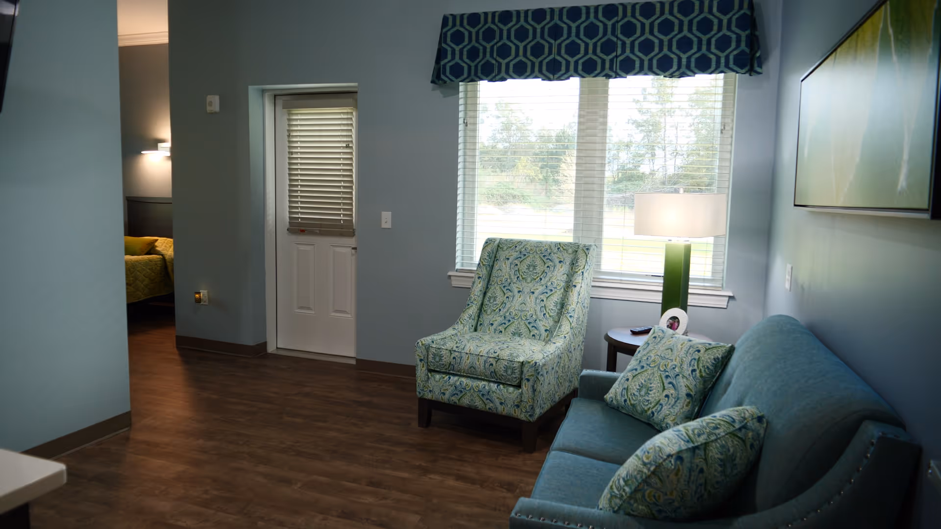 A cozy living room area with a blue patterned armchair and matching sofa with decorative pillows. A side table with a lamp is placed next to the armchair in front of a window with blinds and a blue valance. The room has wooden flooring and light blue walls. A doorway leads to a bedroom with a bed visible in the background.
