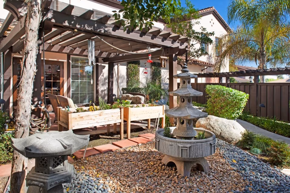 Outdoor garden area at Ivy Park at San Marino featuring a stone pagoda-style water fountain surrounded by pebbles, raised wooden planter boxes with plants, a pergola with hanging wind chimes, and a seating area with cushions. The background shows part of the building exterior with windows and a wooden fence, along with palm trees and other greenery.