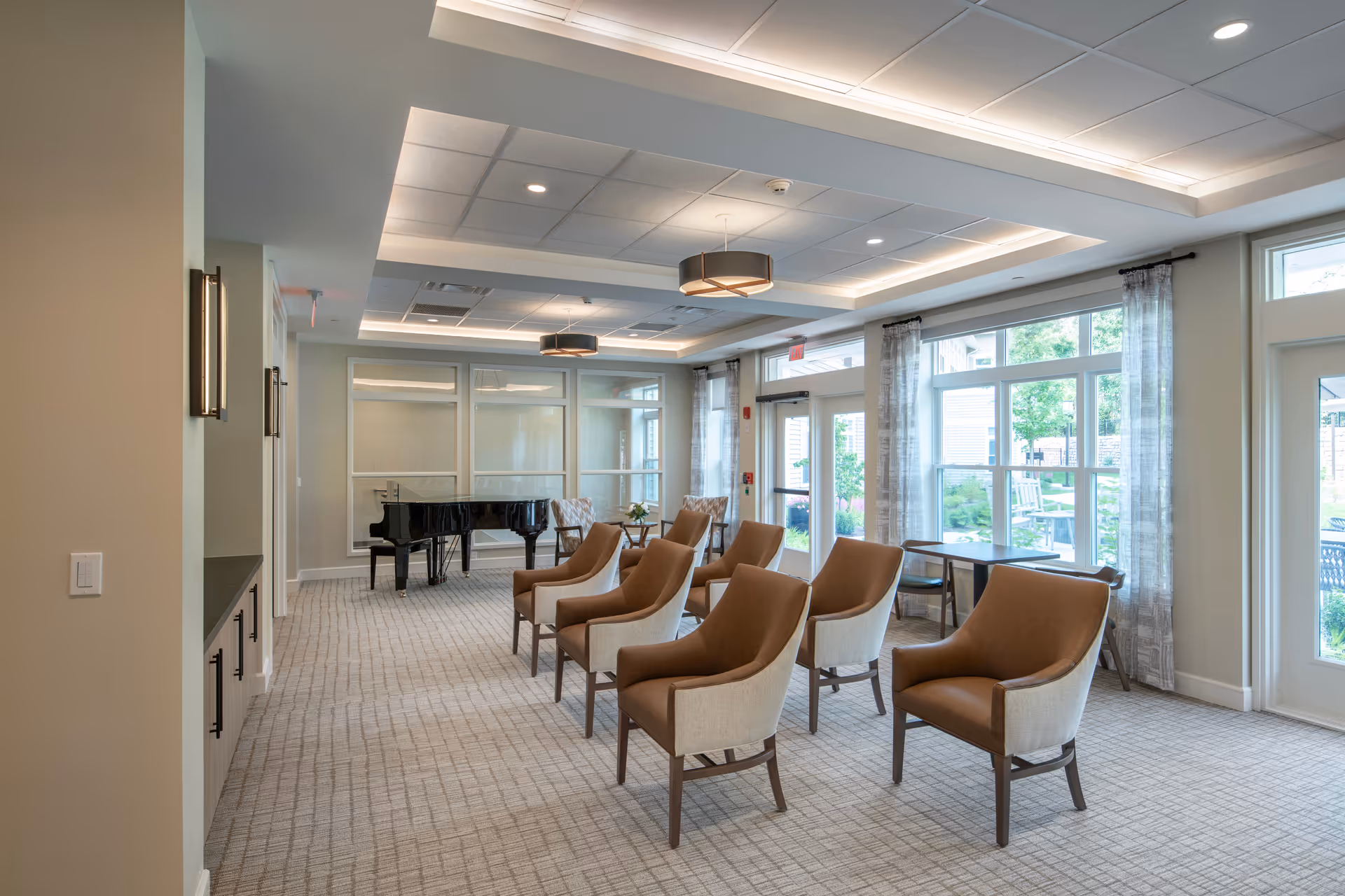 A bright and spacious common area with multiple brown and beige armchairs arranged in rows facing a black grand piano. Large windows with sheer curtains allow natural light to fill the room, and modern ceiling lights provide additional illumination. The room has a neutral color palette with light walls and carpeted floors.