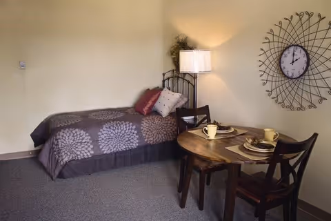 A small senior living facility room featuring a single bed with a dark floral-patterned bedspread and two pillows. Next to the bed is a floor lamp and a small round wooden table set for two with yellow mugs and plates. A decorative wall clock with a wireframe design hangs on the wall above the table.
