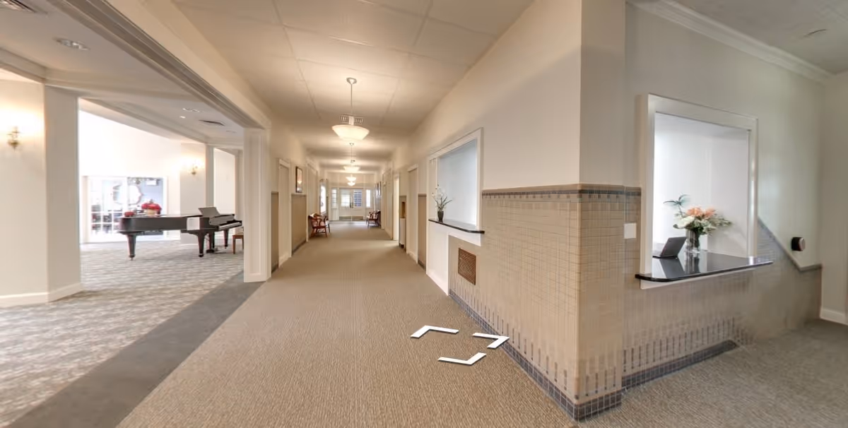 A long, well-lit hallway in a senior living facility with beige walls and carpeted floors. To the left, there is an open room with a grand piano and chairs. On the right side of the hallway, there is a reception window with a small counter and a vase with flowers. The hallway has ceiling lights and chairs placed along the walls.