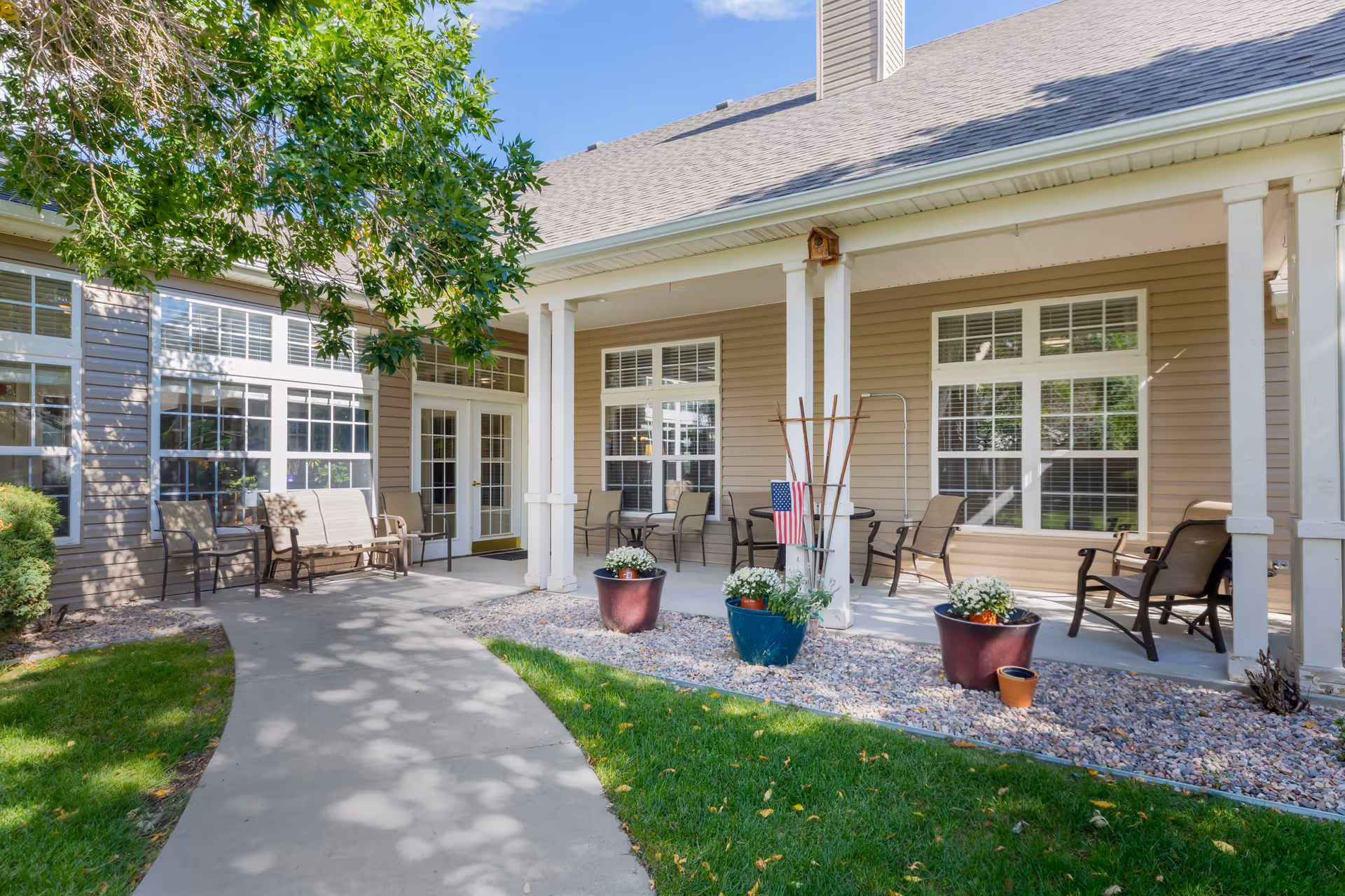 Covered front porch of a senior living building with chairs, potted plants, and a concrete walkway.