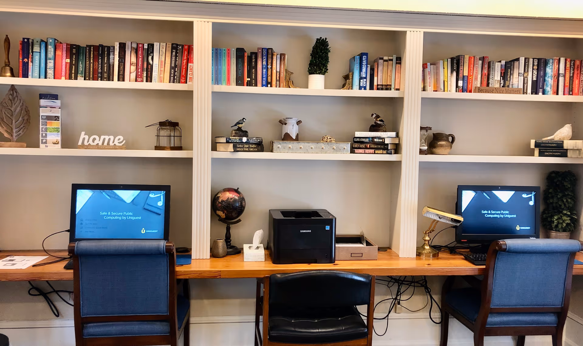 A cozy workspace area with two computer desks and blue chairs in front of built-in white shelves filled with books and decorative items. A printer, globe, tissue box, and desk lamp are placed on the wooden desk. The shelves have various books, small plants, and decorative objects, including a sign that says 'home'.