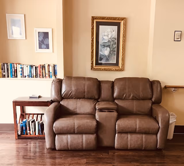 Brown leather reclining loveseat with a center console in a cozy common room, a small bookshelf and framed artwork on the wall behind it.