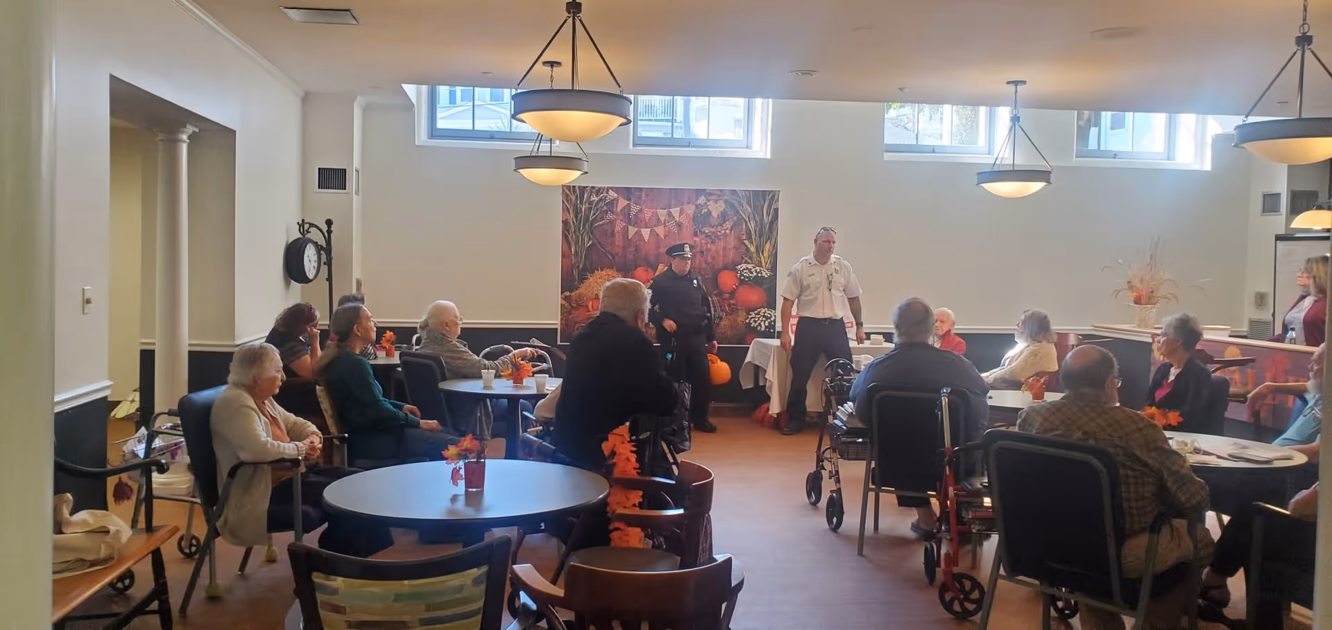 A group of elderly people seated around tables in a well-lit room with large windows near the ceiling. Two men, one in a police uniform and the other in a white shirt, stand at the front of the room addressing the group. The room has light-colored walls, a large decorative painting, and small autumn-themed centerpieces on the tables.