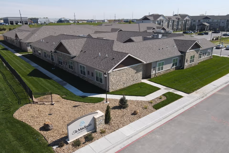 Aerial view of The Madison Senior Living facility showing a single-story building with stone and siding exterior, surrounded by well-maintained green lawns and paved walkways. There is a landscaped area with rocks and a sign displaying the facility's name near the entrance.