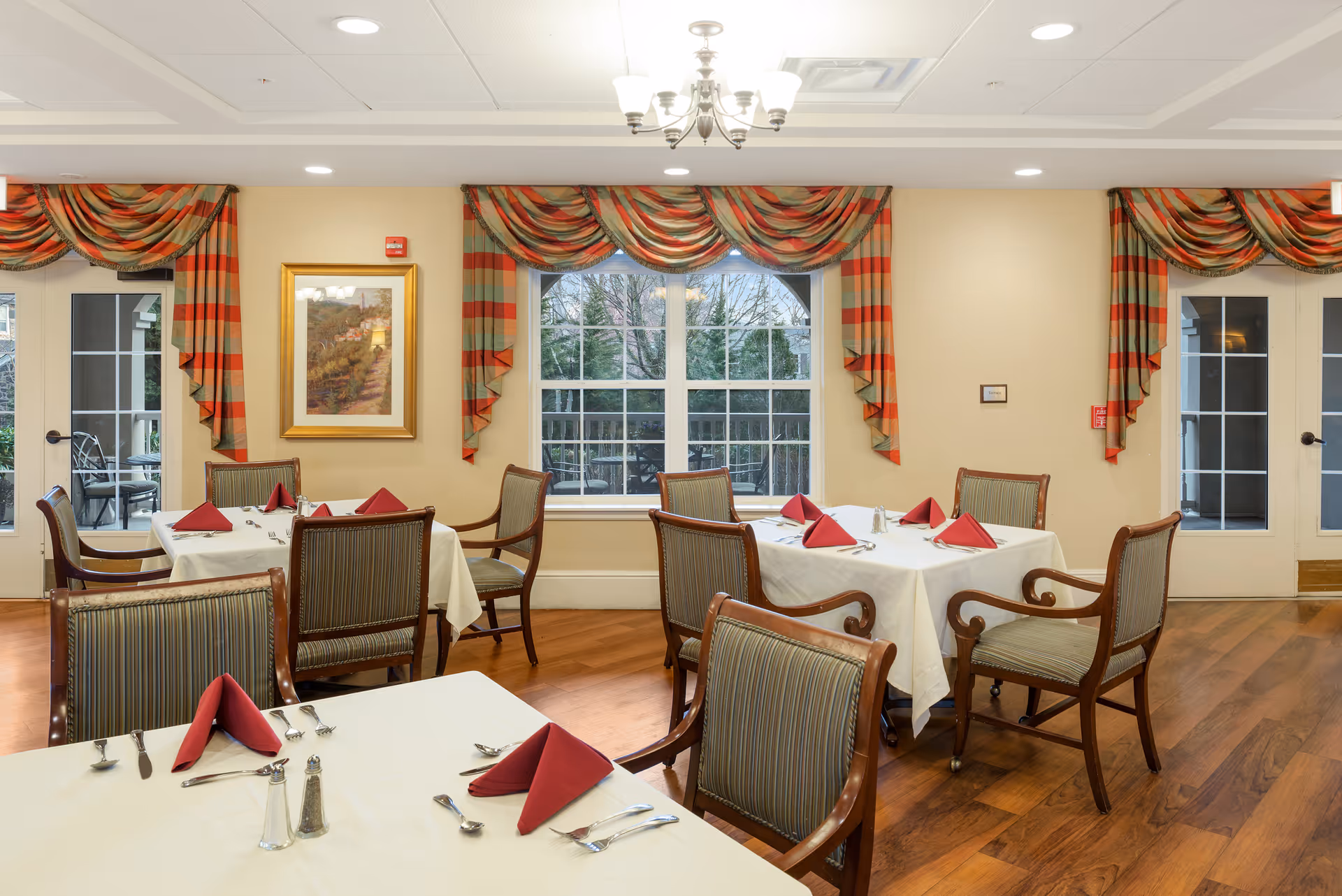 A dining room with several tables covered in white tablecloths and set with red folded napkins, silverware, and salt and pepper shakers. The room has wooden chairs with striped upholstery, hardwood floors, large windows with striped red and green curtains, and a chandelier hanging from the ceiling.