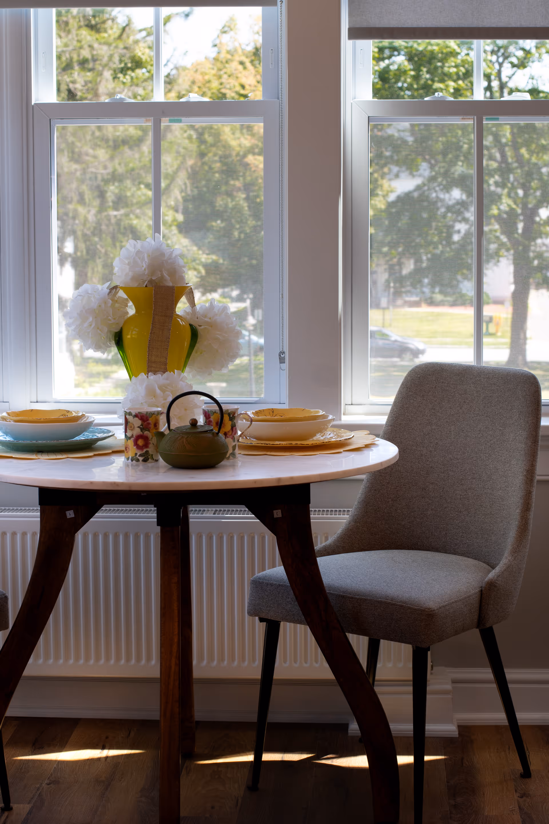 A small round dining table set near two large windows with a view of trees and a street outside. The table has a green teapot, two floral mugs, a yellow vase with white flowers, and two place settings with yellow plates and bowls. A gray upholstered chair is positioned next to the table.