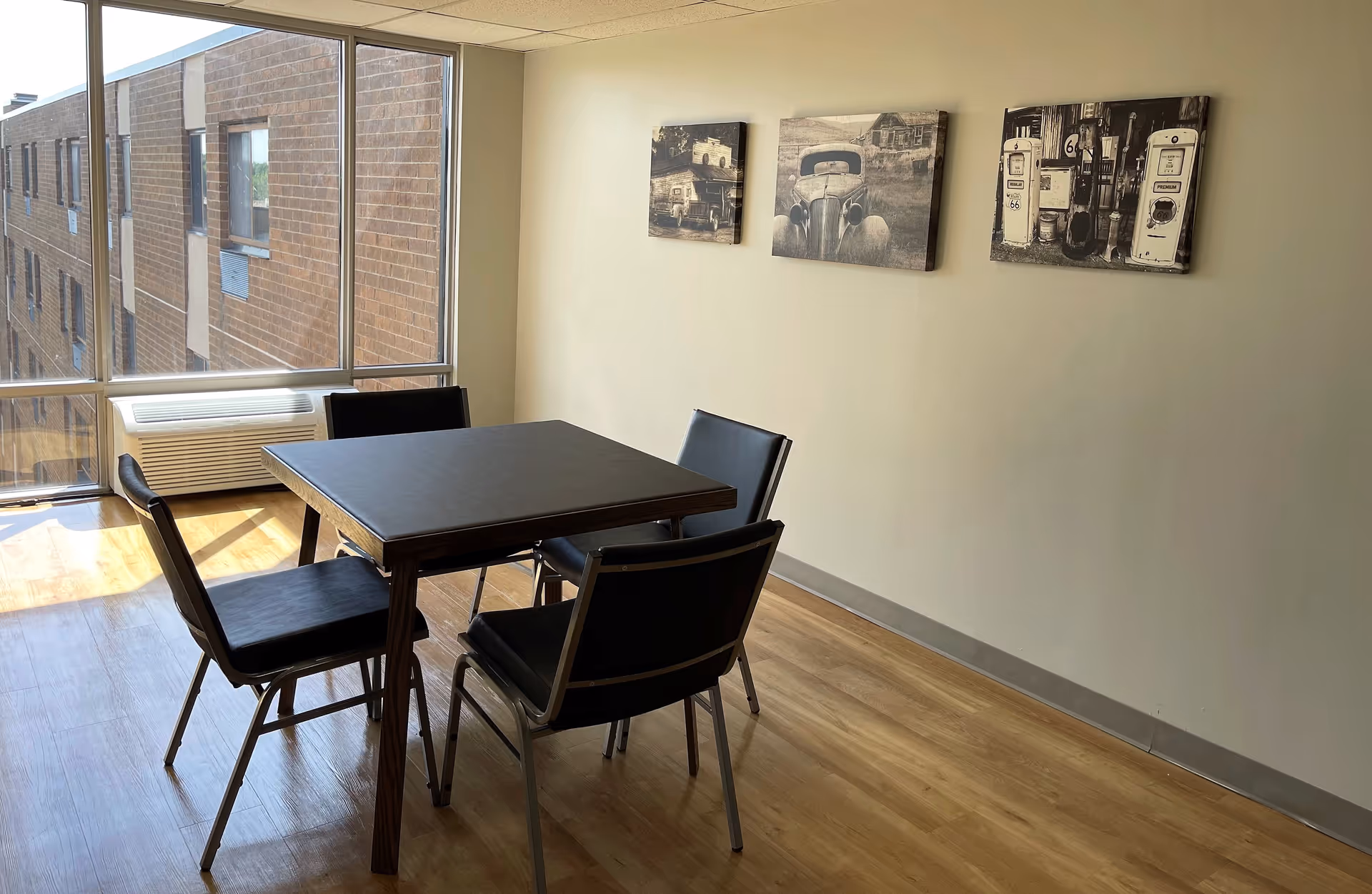 A small dining area with a square table and four black chairs on a wooden floor. The room has large windows letting in natural light and three vintage-style black and white pictures hanging on a beige wall.