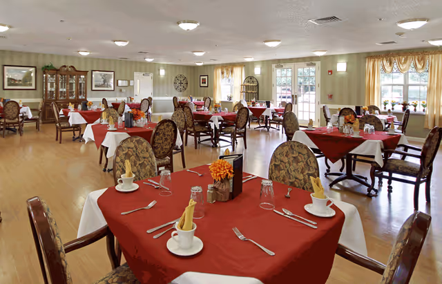 A spacious dining room with multiple tables covered in red and white tablecloths, each set with cups, utensils, glasses, and napkins. The room has wooden flooring, patterned upholstered chairs, large windows with curtains, and soft lighting from ceiling fixtures. Decorative elements include framed pictures on the walls and a wooden cabinet.