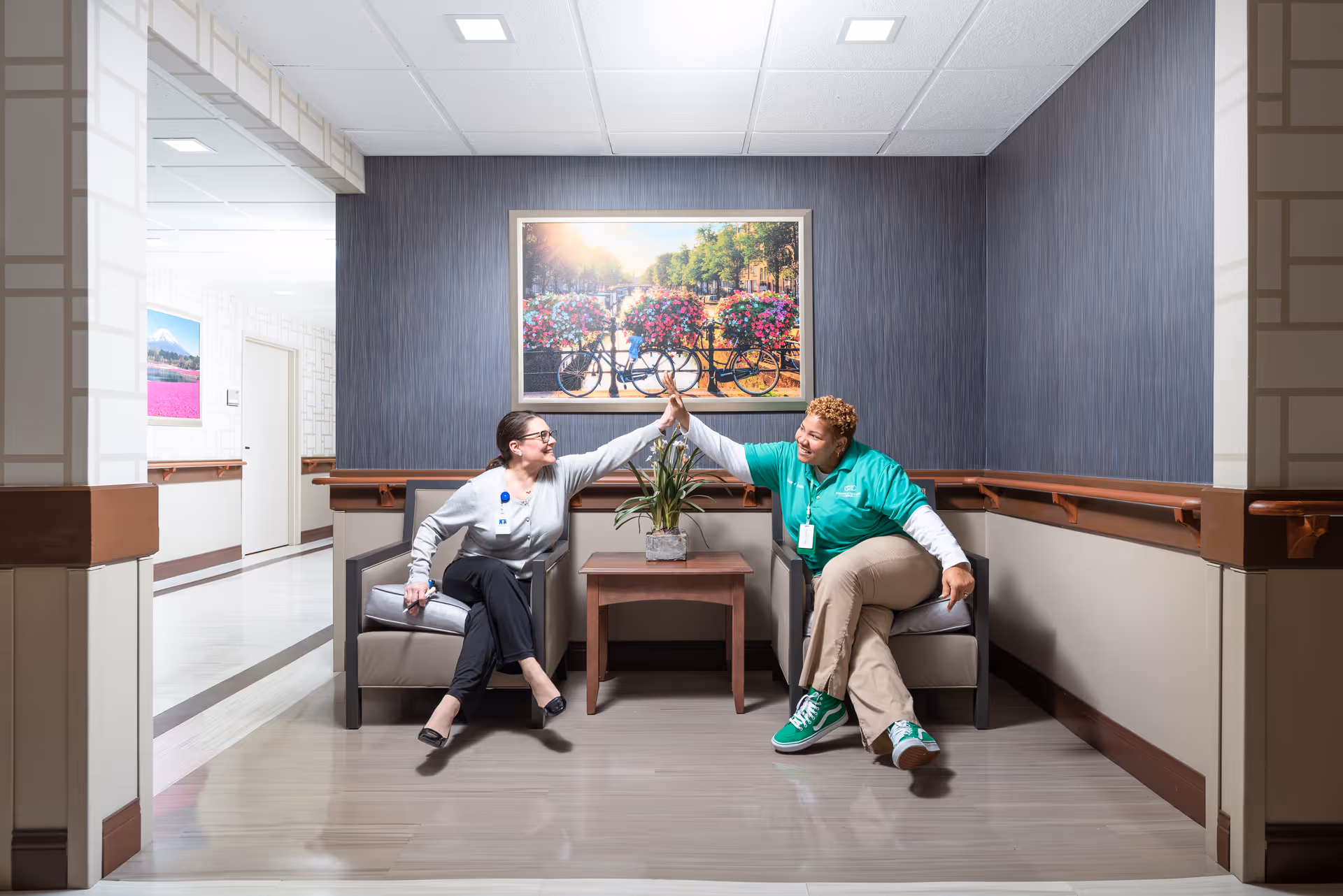 Two women sitting in armchairs in a hallway or common area of a senior living facility, smiling and giving each other a high five. There is a small wooden table with a plant between them, and a colorful framed picture of bicycles with flowers on the wall behind them.