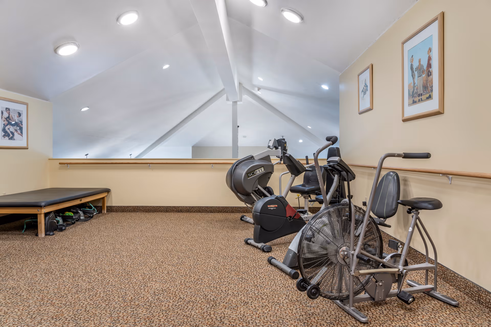 A small exercise room with beige walls and carpeted floor. The room contains several pieces of exercise equipment including a stationary bike, a recumbent bike, and an elliptical machine. There is a padded bench along one wall with some exercise bands or straps underneath it. The ceiling is vaulted with recessed lighting, and three framed pictures hang on the walls.