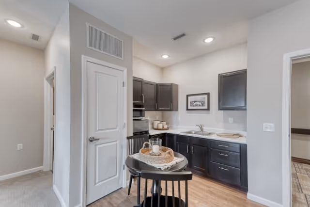 A small kitchen area with dark cabinets, a white countertop, and a stainless steel microwave. There is a round black table with two black chairs in front of the kitchen. On the table is a woven tray holding a candle and some decorative items. The walls are light-colored, and there is a framed picture hanging above the sink. The floor is wood, and there is a white door to the left and an open doorway to the right.