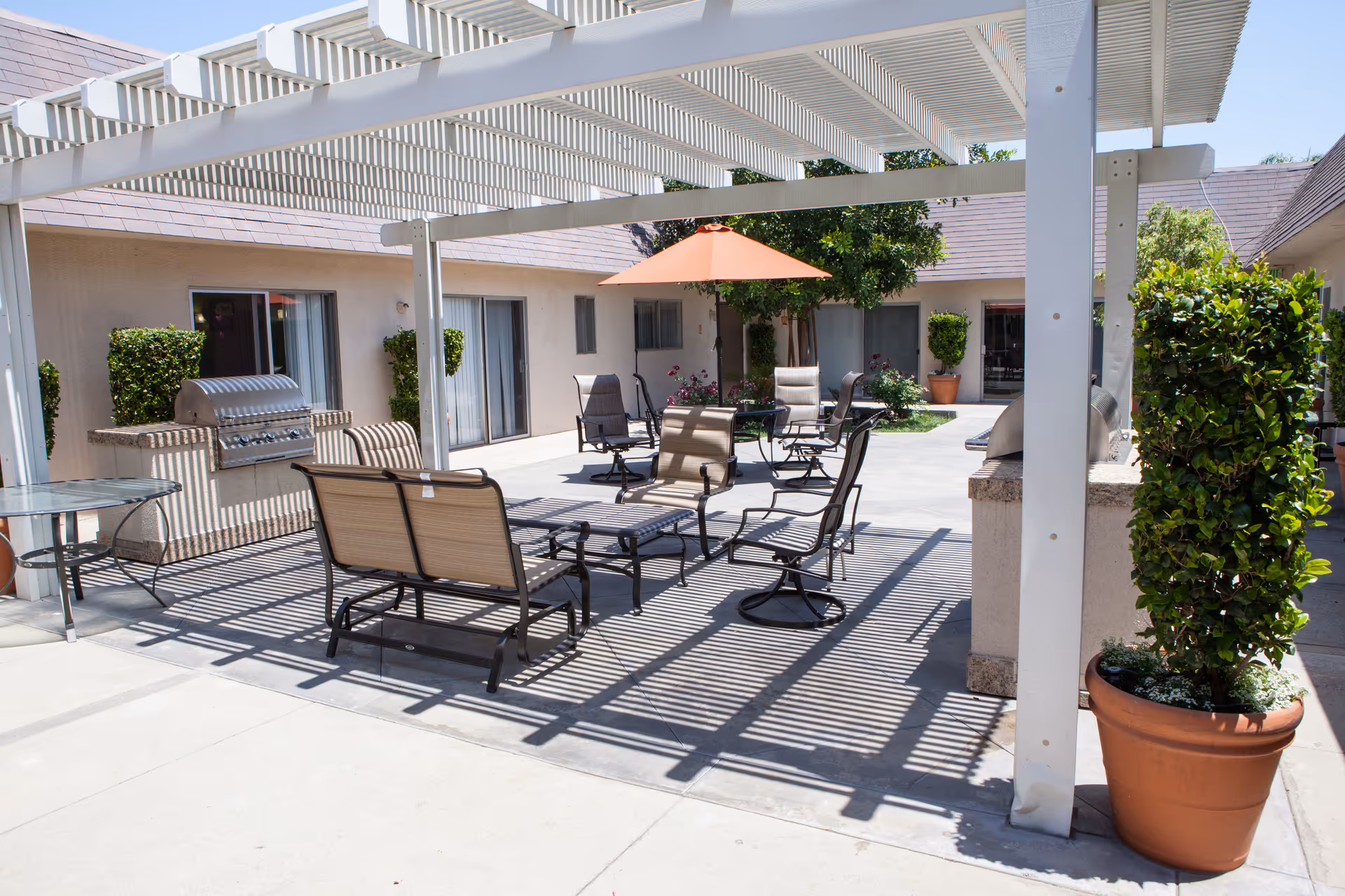Outdoor patio area at Citrus Nursing Center with a white pergola casting shadows over several chairs and tables. There is a built-in grill on the left side and potted plants around the space. An orange umbrella provides shade to some seating in the background, and the patio is surrounded by the building with sliding glass doors and windows.