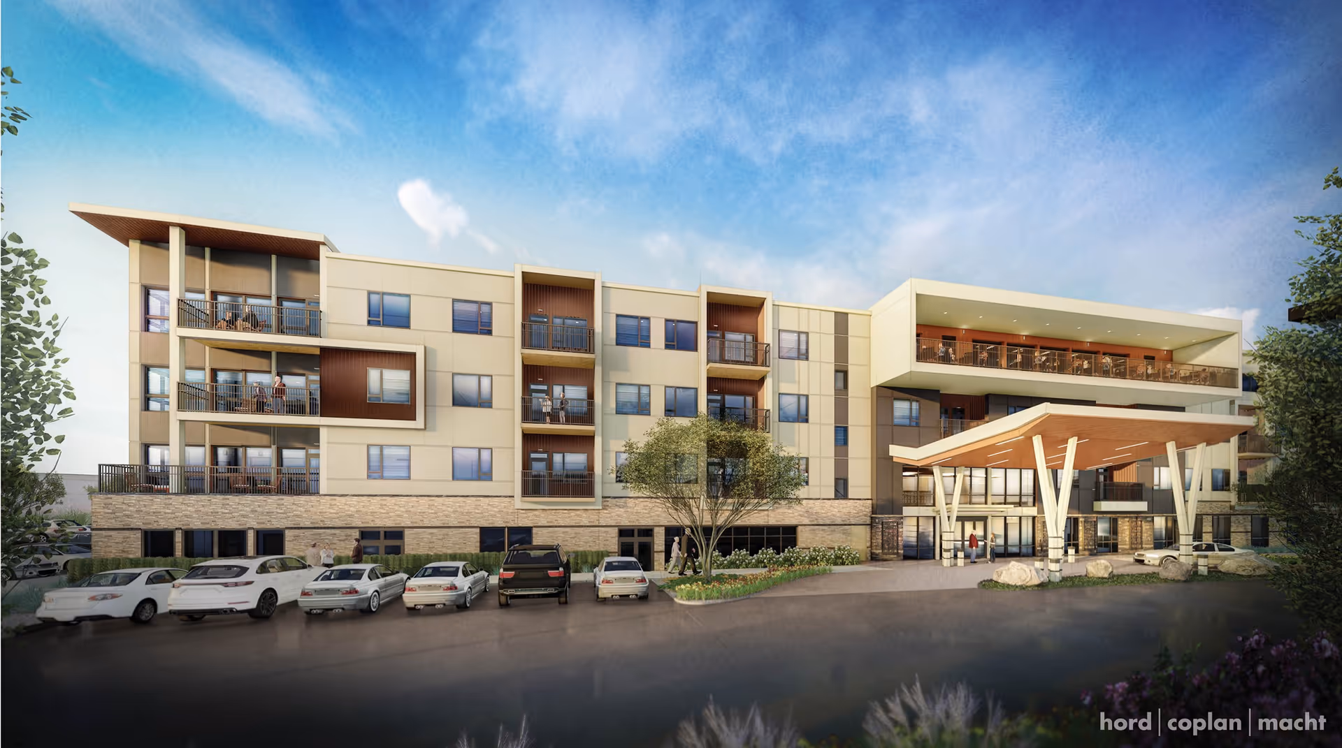 Four-story senior living building with balconies, a covered entrance canopy, landscaping and parked cars under a blue sky.