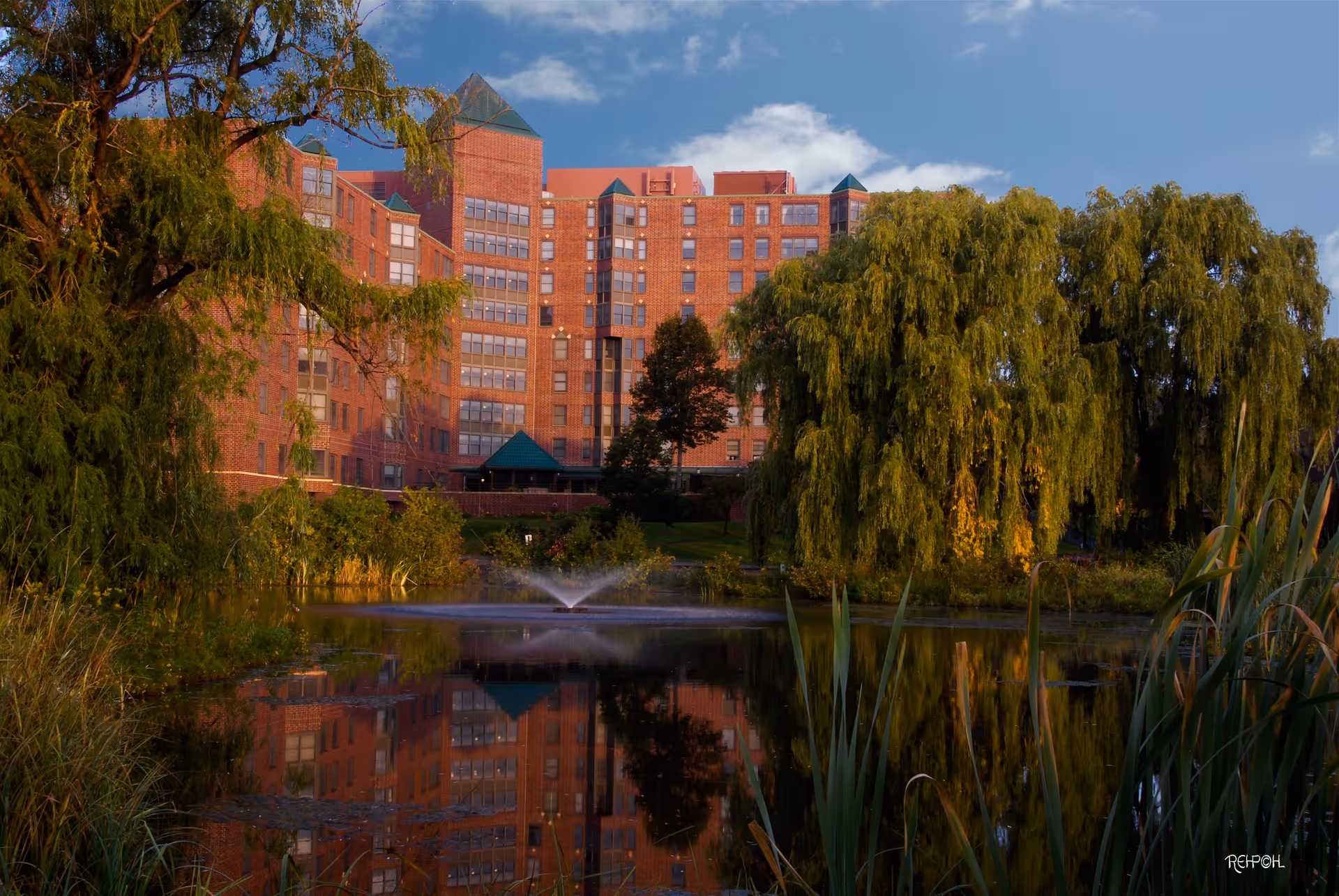 A large brick building with multiple windows and green peaked roofs is reflected in a pond with a water fountain in the center. The pond is surrounded by lush greenery, including tall trees and reeds, under a partly cloudy blue sky.