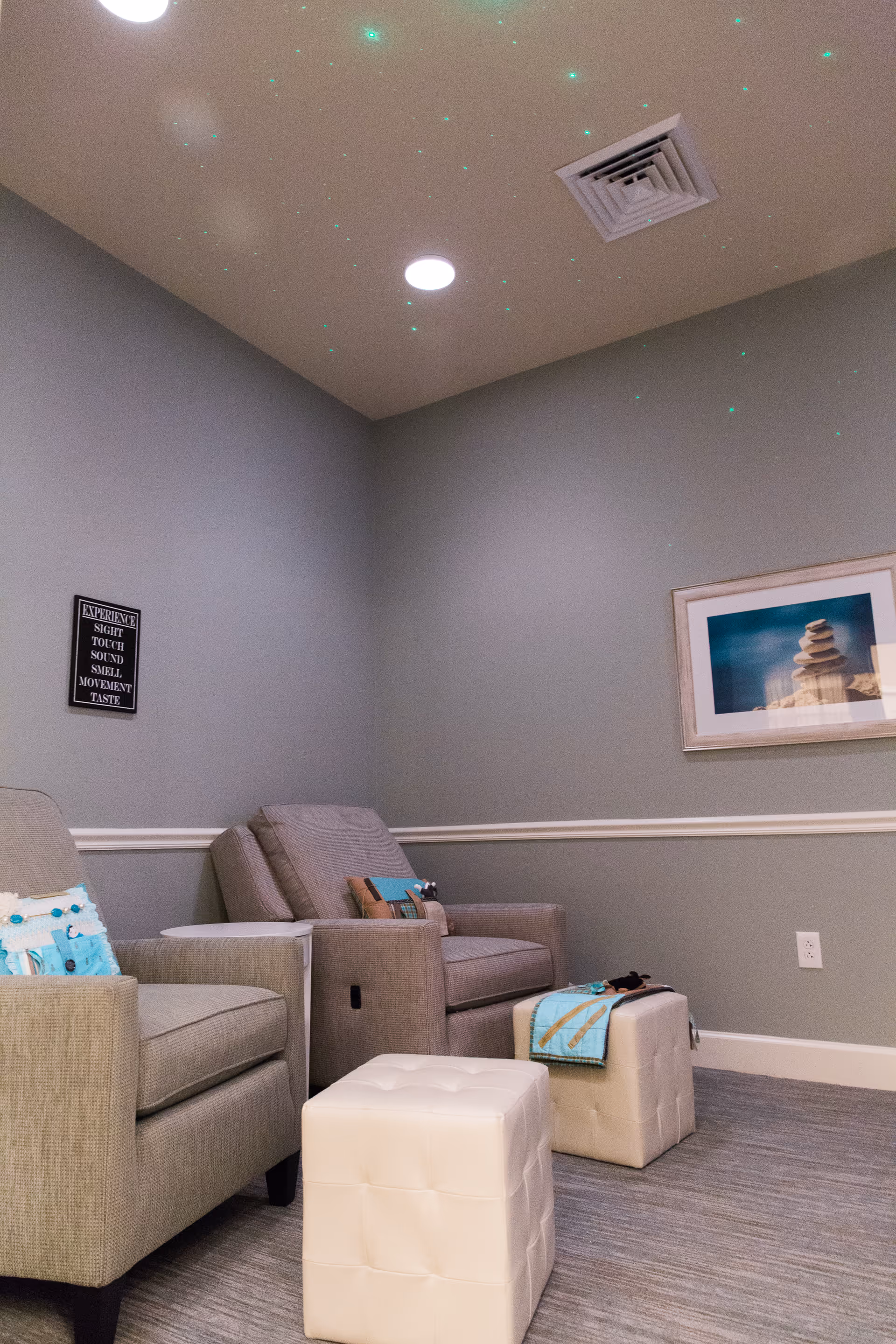 A cozy corner of a room with two gray armchairs, two white cushioned ottomans, and a small white side table. The walls are painted gray with white trim, and there is a framed picture of stacked stones on the wall. A small black sign with white text is also visible on the wall. The ceiling has a vent and a round light fixture, with small green star-like lights projected onto it.