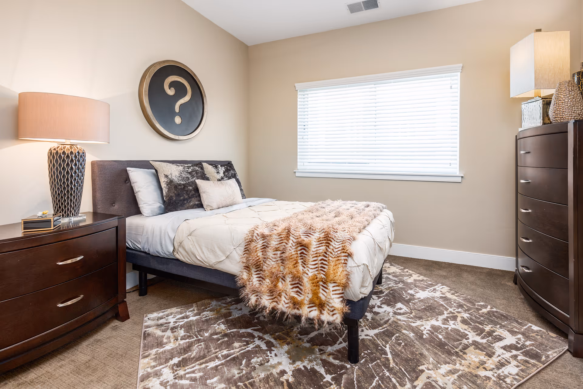 A neatly made bed with a beige comforter and a furry throw blanket in a bedroom. The bed has multiple pillows and is flanked by a dark wooden nightstand with a decorative lamp on the left and a matching dark wooden dresser with a lamp and decorative items on the right. A large window with white blinds is on the wall behind the bed, and a round wall decoration with a question mark hangs above the headboard.