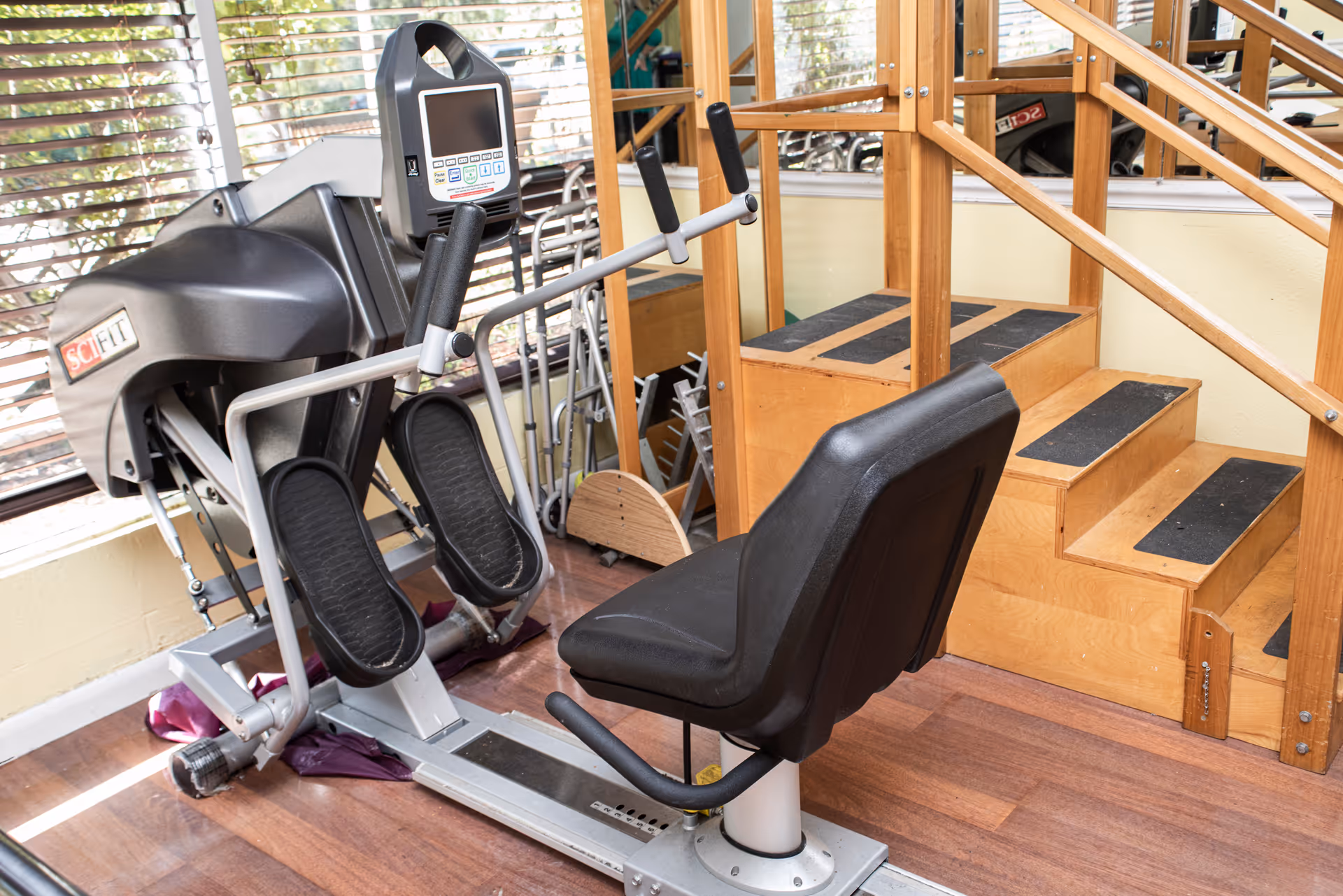 A small rehabilitation gym area with exercise equipment, a seated machine, and wooden therapy stairs.