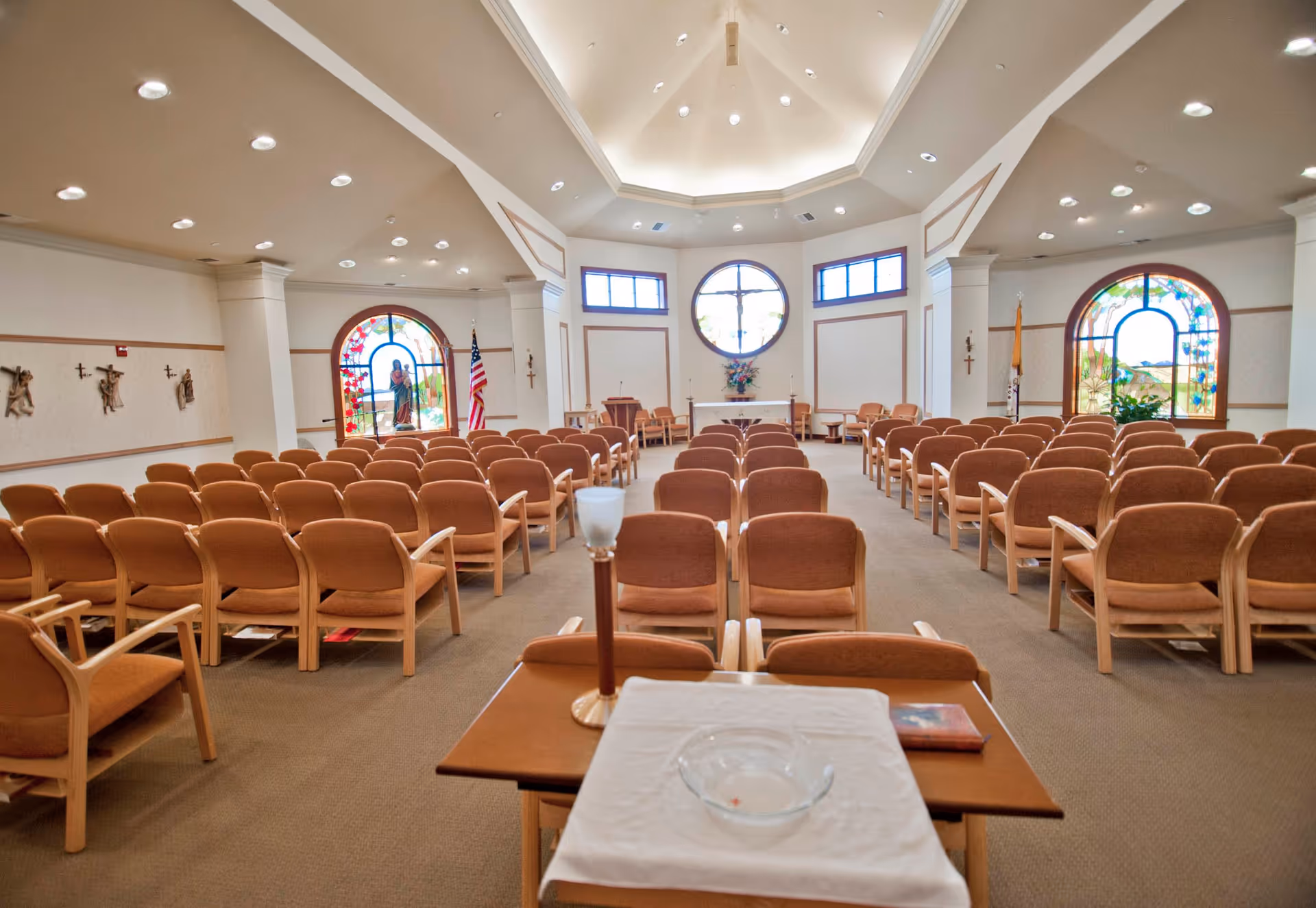 Interior view of a chapel with rows of brown cushioned chairs facing an altar. The chapel features stained glass windows, religious statues, crosses on the walls, and a high ceiling with recessed lighting.