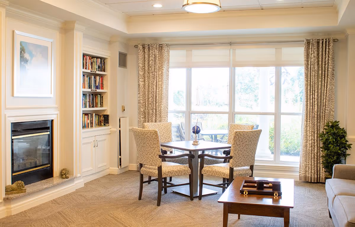 A bright and cozy living room area with a fireplace on the left, built-in bookshelves, a square table with four upholstered chairs in front of large windows with patterned curtains, and a coffee table with a tray on it. There is also a sofa partially visible on the right and a potted plant near the window.