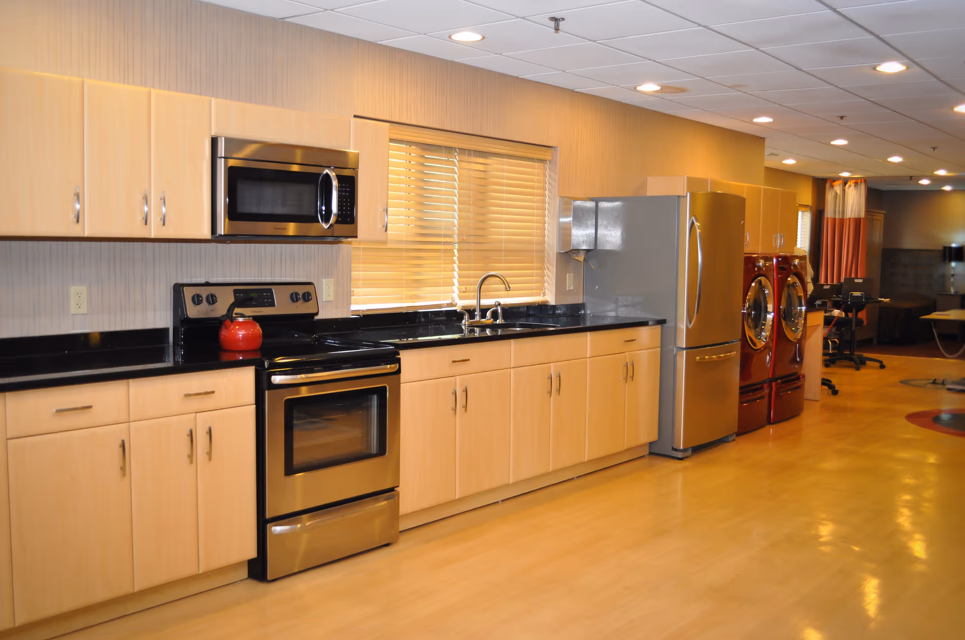 A modern kitchen area with light wood cabinets, a stainless steel microwave, stove, and refrigerator. There is a red kettle on the stove and a window with blinds above the sink. To the right, there are two red front-loading washing machines and dryers, and an office chair and desk are visible in the background.