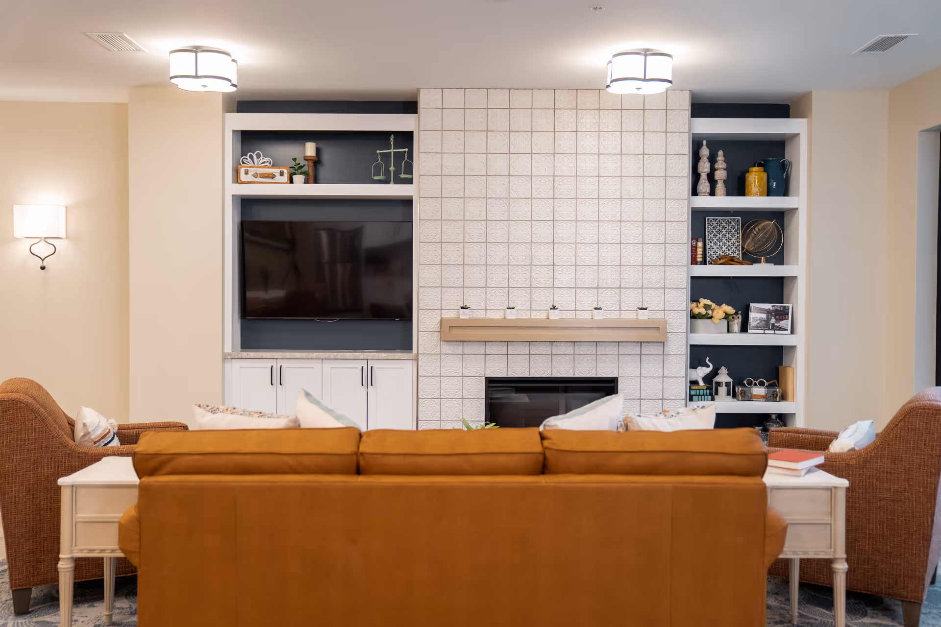 A cozy living room area featuring a brown leather sofa facing a tiled fireplace with a wooden mantel holding small decorative items. On either side of the fireplace are built-in shelves with various decorations and a flat-screen TV on the left. Two wicker armchairs flank the sofa, and side tables with books and pillows complete the seating arrangement. The room is softly lit with ceiling and wall lights.