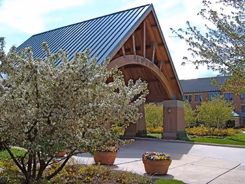 Entrance area of a senior living facility with a large covered driveway supported by brick pillars and a metal roof. Flowering trees and potted plants with colorful flowers surround the paved driveway. A multi-story brick building is visible in the background under a clear blue sky.