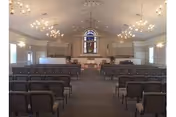 Interior view of a chapel-style assembly room with rows of chairs facing a raised platform and a stained-glass window.
