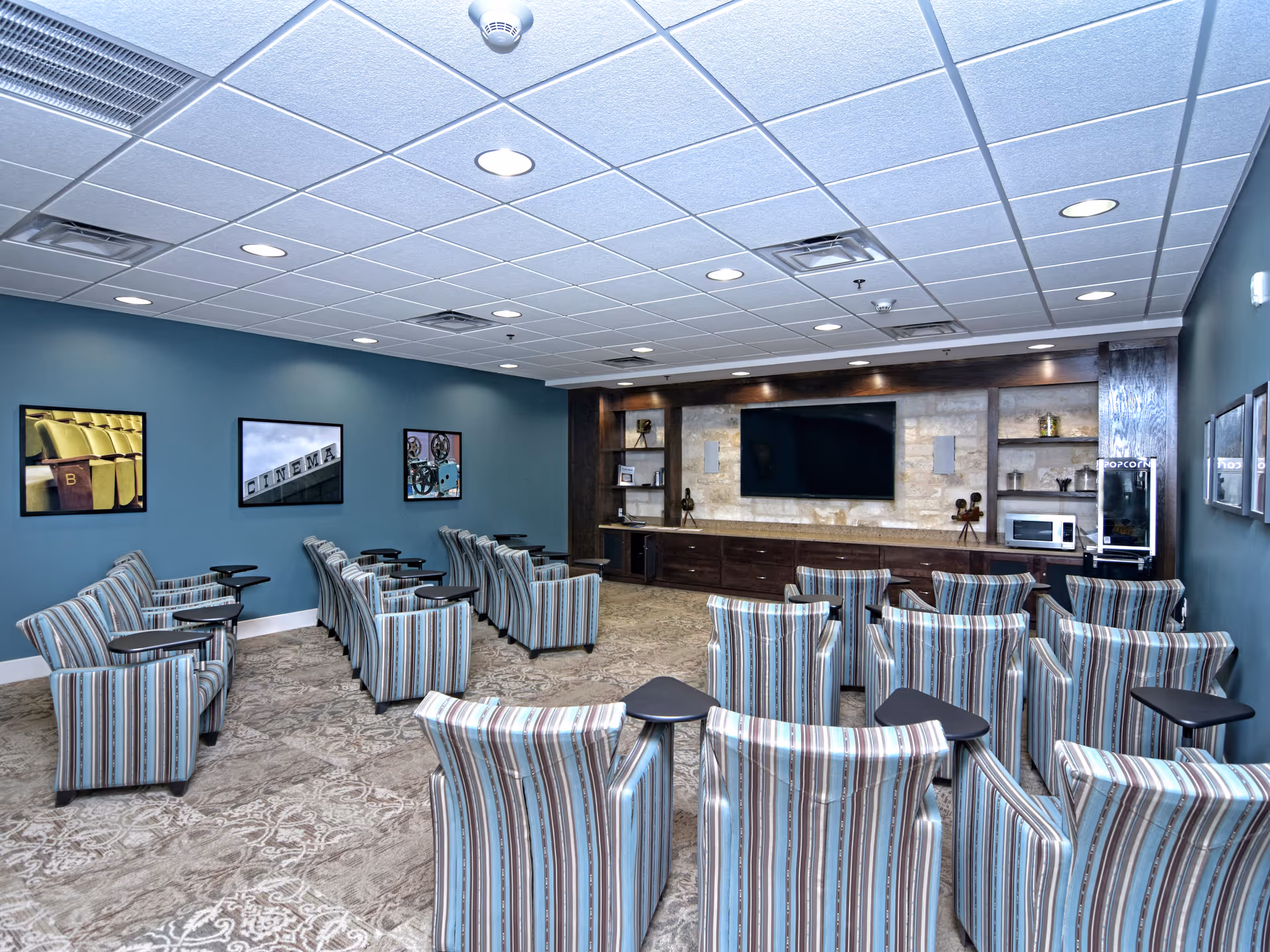 A small theater room with multiple striped armchairs arranged in rows facing a large flat-screen TV mounted on a stone wall. The room has blue walls adorned with framed pictures related to cinema, a popcorn machine, and a microwave on a wooden built-in shelf unit.