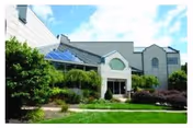 Exterior view of a modern senior living facility building with large windows, a glass entrance canopy, and surrounding landscaped greenery under a blue sky with some clouds.