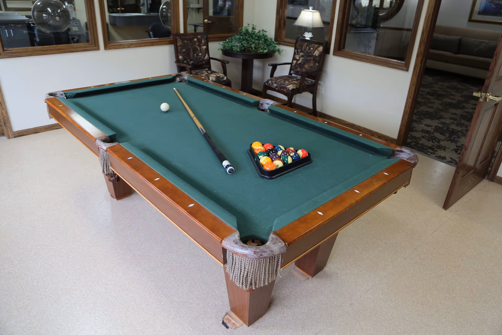A pool table with a green felt surface in a room. The table has a cue stick, a white cue ball, and a rack of colorful billiard balls arranged in a triangle. Behind the table are two wooden chairs with patterned cushions, a small round table with a green plant, and a lamp. The room has beige flooring, white walls, and several large windows with wooden frames. An open door reveals another room with a patterned carpet and a sofa.