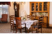 A traditional dining room with a round table covered by a white lace tablecloth, surrounded by six wooden chairs. In the background, there is a wooden china cabinet with glass doors displaying glassware and decorative items. A grandfather clock stands near a window with sheer curtains and a valance. The room has patterned carpet and wallpaper with vertical stripes.