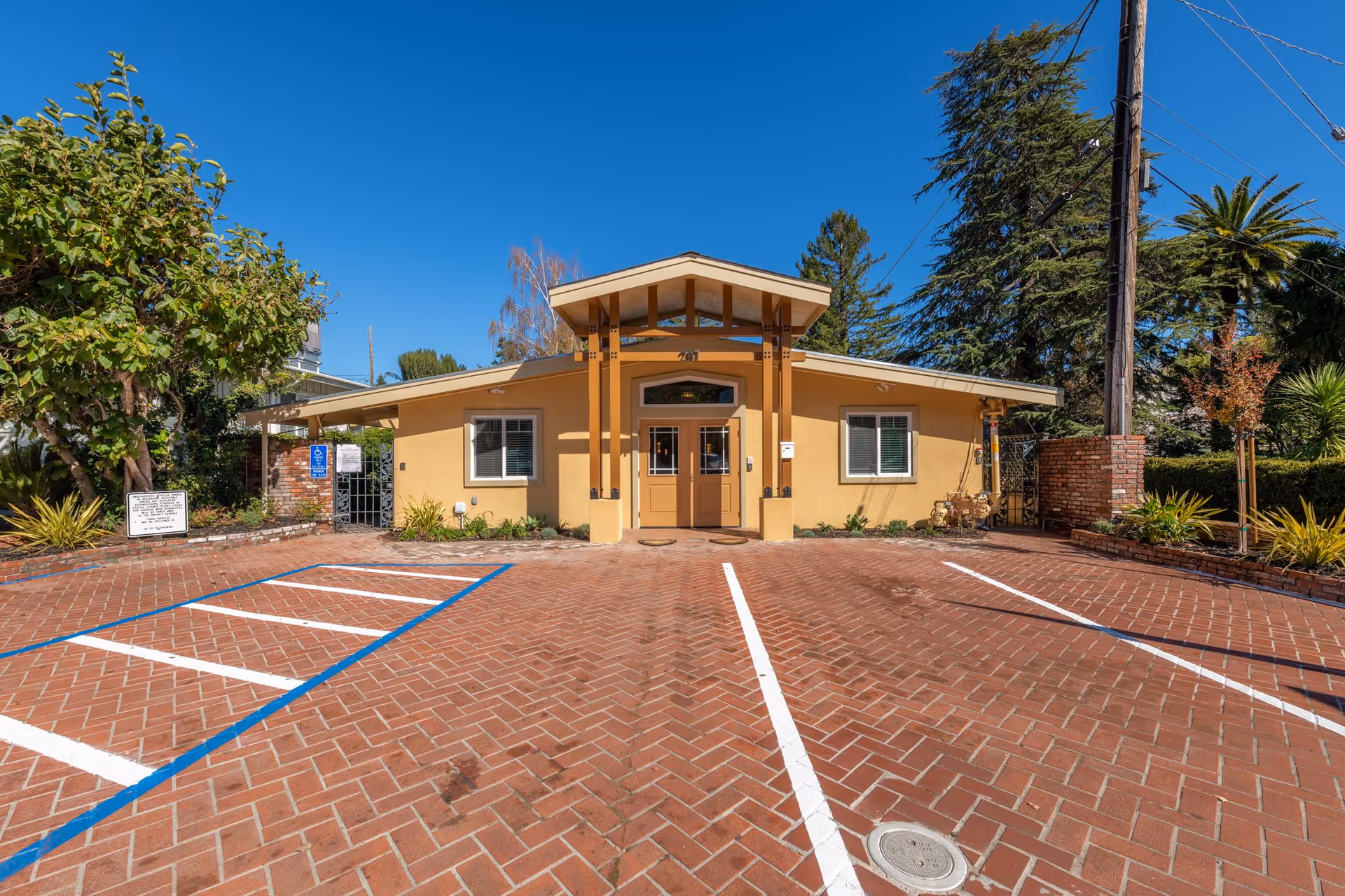 Front exterior view of a single-story building with a tan facade and a covered entrance supported by wooden beams. The building is surrounded by trees and plants, with a brick-paved parking area in front, including a handicapped parking space. The sky is clear and blue.
