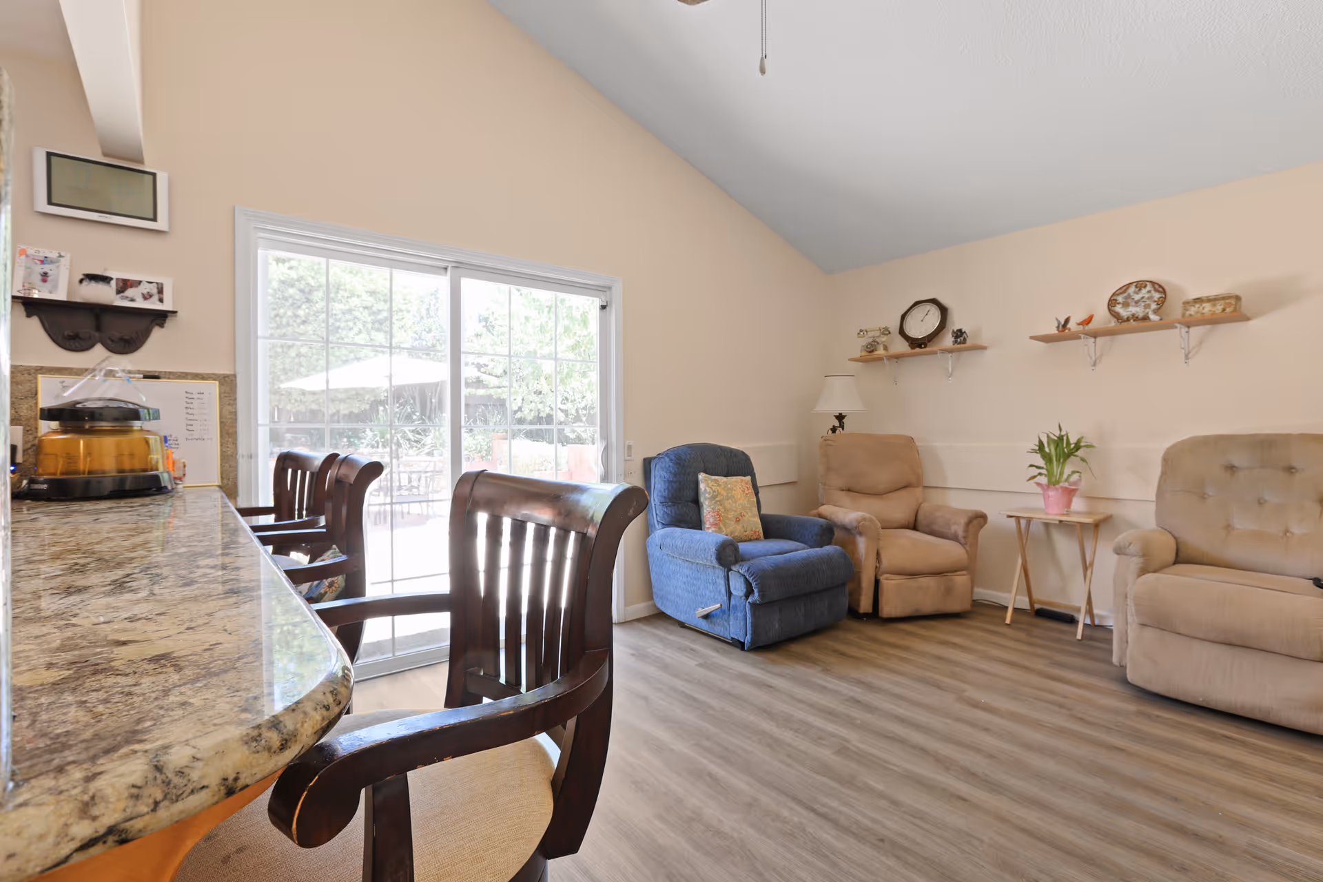 A cozy living room area with three armchairs in blue and beige colors, a small side table with a potted plant, and two wall shelves displaying decorative items. To the left, there is a granite countertop with wooden chairs and a sliding glass door leading to an outdoor patio with an umbrella and greenery.