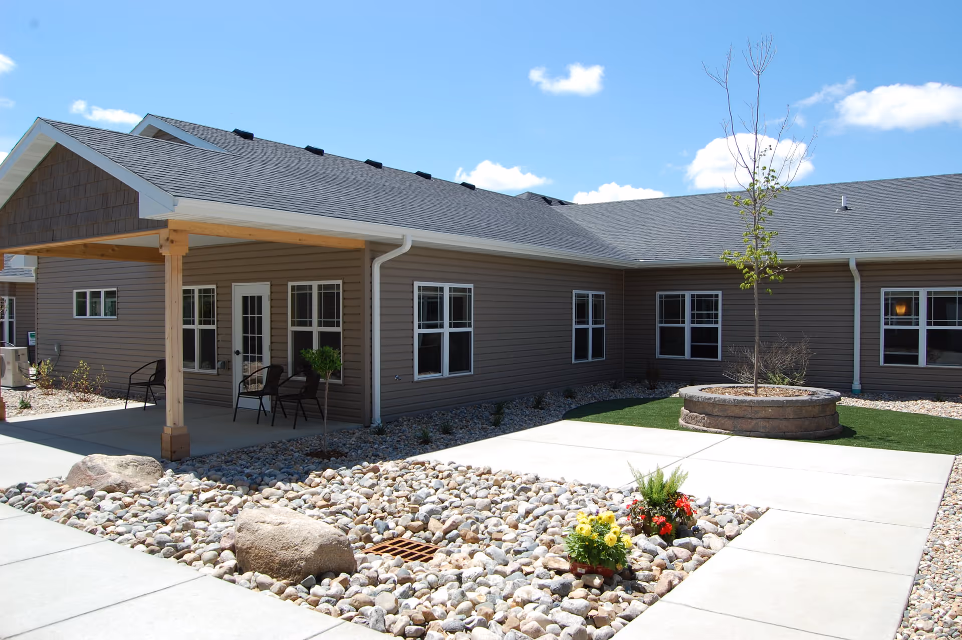 Exterior view of a single-story assisted living facility building with beige siding, multiple windows, and a covered porch area with chairs. The courtyard features a rock garden with large stones, a small flower bed, a young tree planted in a circular stone planter, and concrete walkways under a bright blue sky with scattered clouds.