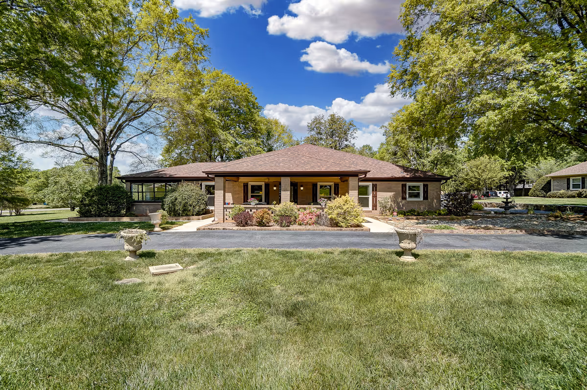 Single-story brick building with a covered entrance surrounded by lawn, trees, and planters under a blue sky.