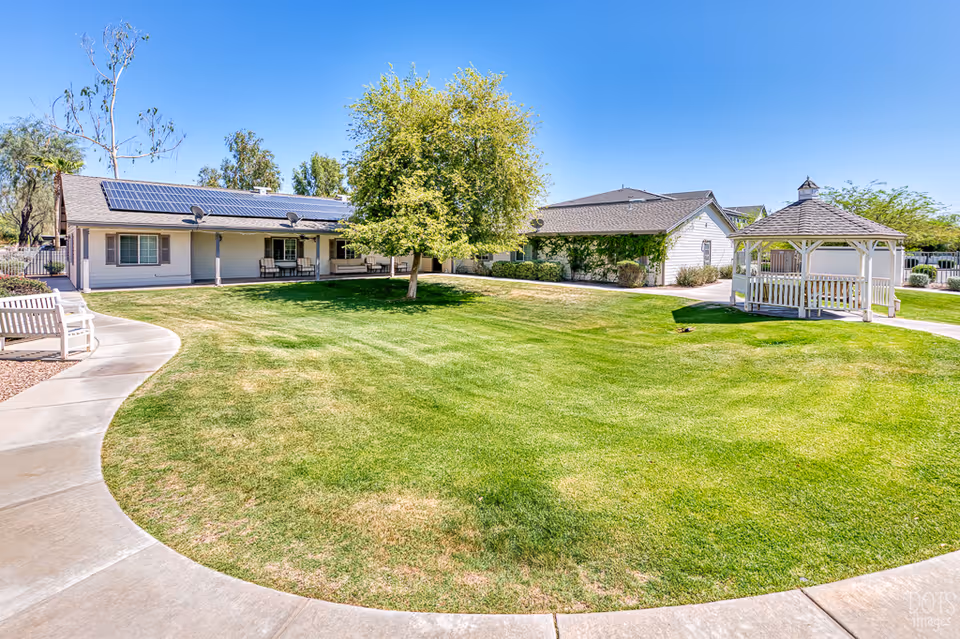 A sunny outdoor area at Paradise Valley Senior Living featuring a well-maintained green lawn, a large tree in the center, a white gazebo on the right, and a curved concrete walkway. There are benches along the walkway and single-story buildings with solar panels on the roof in the background under a clear blue sky.