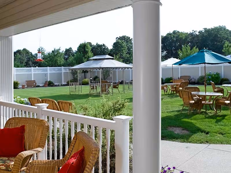 Covered porch with wicker chairs overlooking a grassy courtyard with tables, umbrellas, and a gazebo.