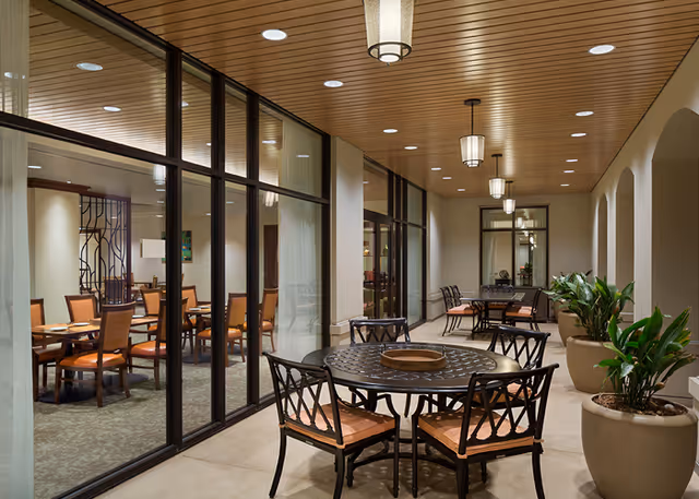 Covered outdoor seating area at MonteCedro Retirement Community with round metal tables and cushioned chairs, large potted plants, and ceiling lights. Glass walls separate this area from an indoor dining room with wooden chairs and tables.