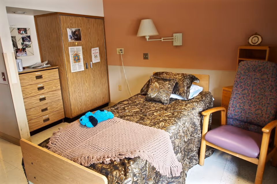 A single bed in a senior living room with a patterned bedspread, pillows, a pink throw and a small blue stuffed toy, flanked by a wooden wardrobe, dresser and an upholstered armchair.